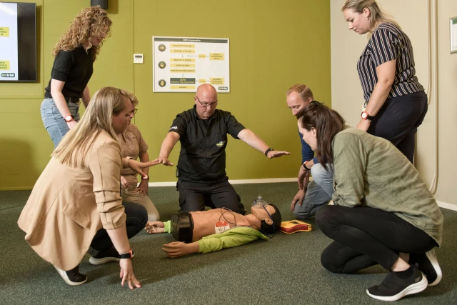 Groep mensen kijkt naar de EHBO trainer die aangeeft dat de pop niet aangeraakt mag worden